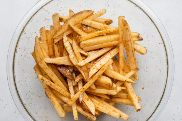 Overhead view of uncooked potato sticks tossed in cajun seasoning and avocado oil in a large glass mixing bowl.