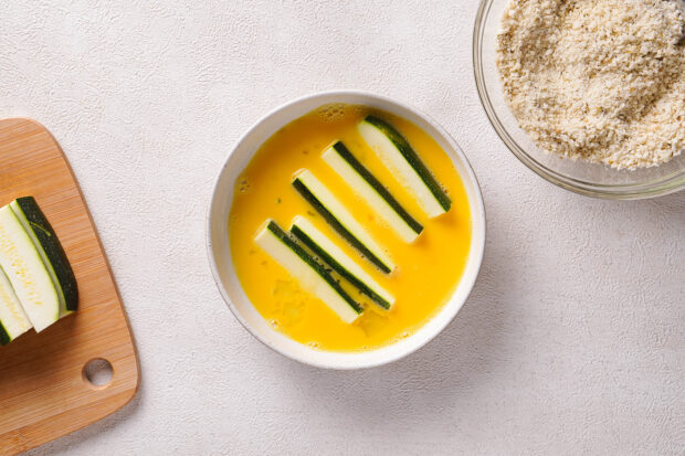 Overhead view of zucchini sticks in egg wash in small ceramic white bowl.