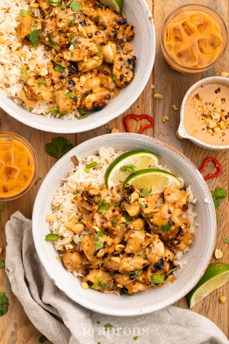 Zoomed out, overhead view of 2 bowls of Thai peanut chicken and white rice with lime wedges on a wooden table.