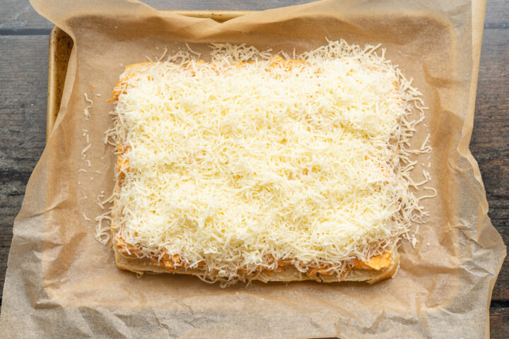 Overhead view of buffalo chicken topped with mozzarella, spread out on top of a slab of halved sweet dinner rolls in a baking dish lined with parchment paper.