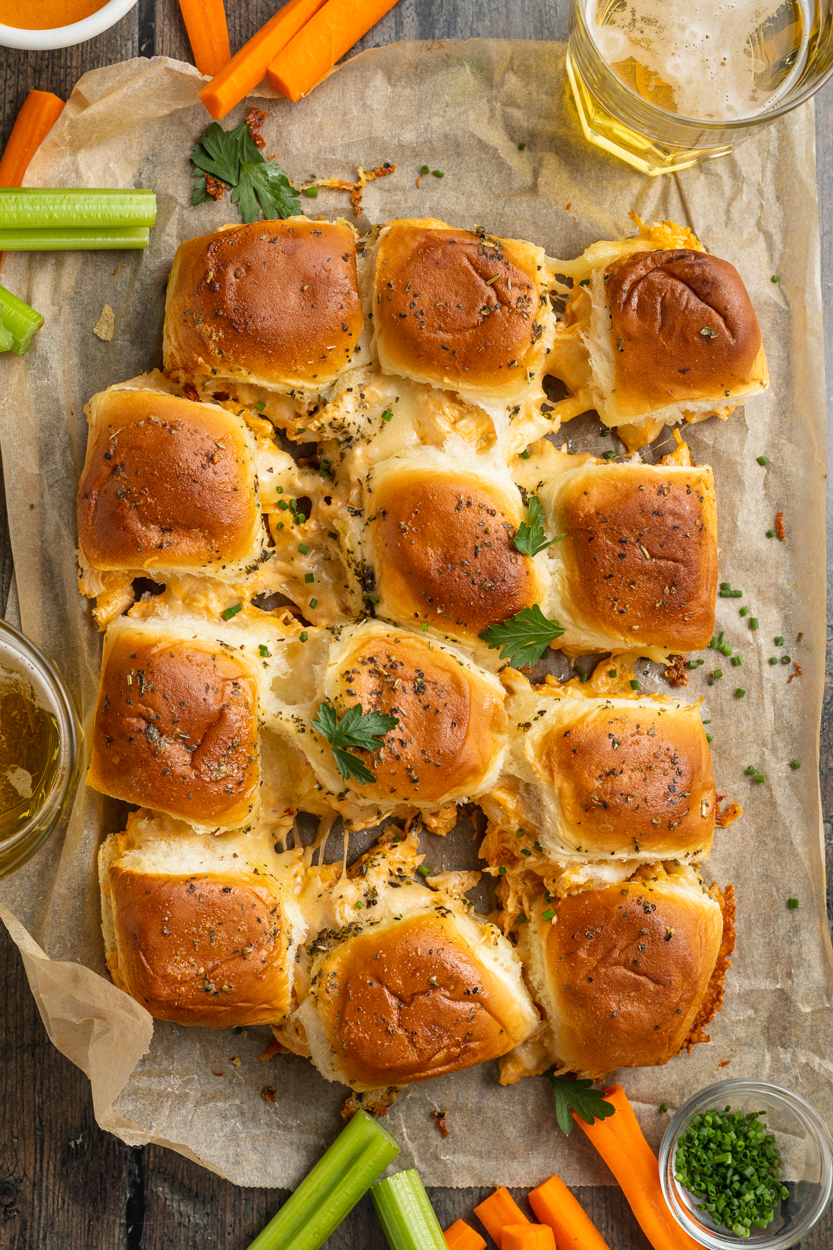 Zoomed out, overhead view of one dozen buffalo chicken sliders arranged in 4 rows of 3 on a wooden table.