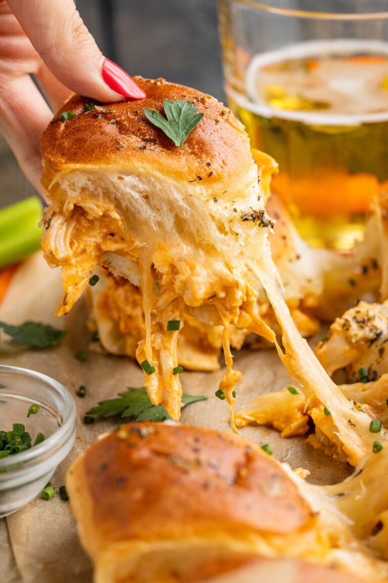 A close, angled view of a white woman's hand pulling a buffalo chicken slider away from other sliders, with an ooey gooey cheese pull connecting them.