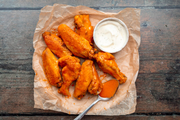 Overhead view of orange, saucy sous vide chicken wings on a round plate lined with parchment paper, with a small ramekin of ranch dressing.