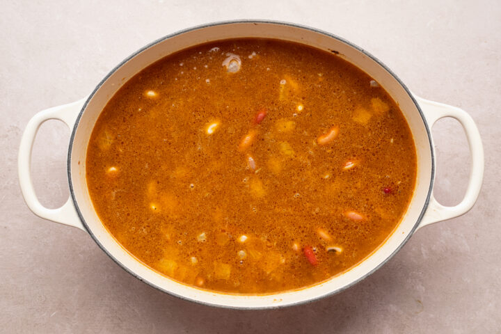 Overhead view of chicken broth, onion, celery, and a variety of spices for 15 bean soup in a large oval ceramic pot with 2 handles.