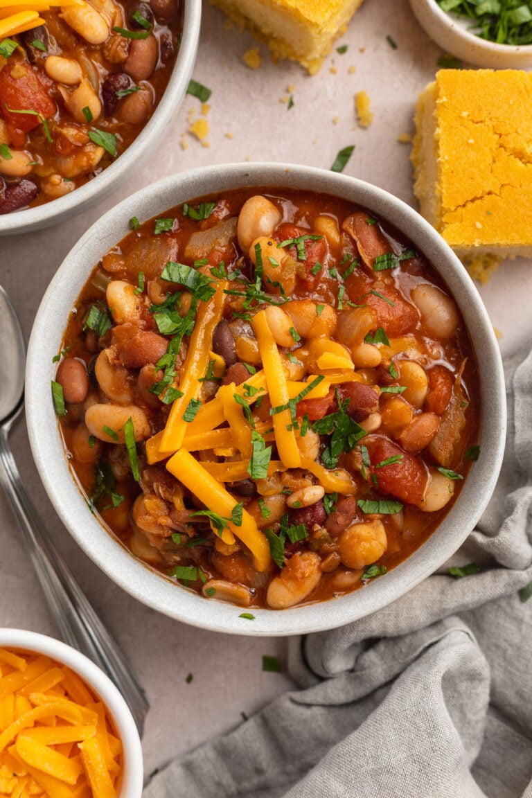 Overhead view of a bowl of deep red 15 bean soup topped with chopped green onions and yellow shredded cheddar in a white bowl on a white table with spoons, chopped onions, and cornbread.