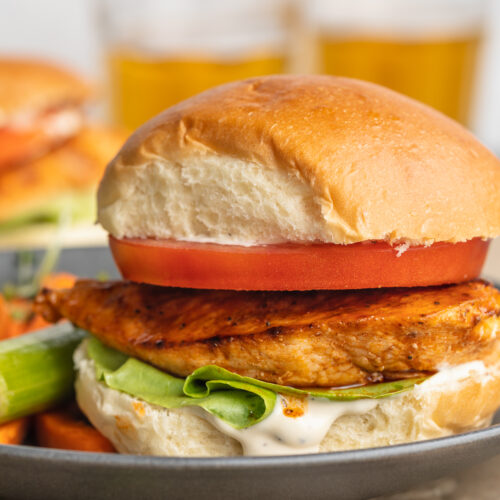 Side view of a buffalo chicken sandwich with an orange-colored grilled chicken breast, a thick slice of tomato, green lettuce, and a fluffy bun on a blue-grey plate.