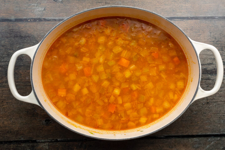 Overhead view of buffalo sauce, chicken broth, and shredded chicken in a large soup pot on a wooden table.