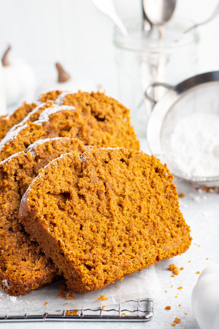 Side view of bright orange slices of gluten-free pumpkin bread topped with powdered sugar leaning up on one another on a white tabletop.