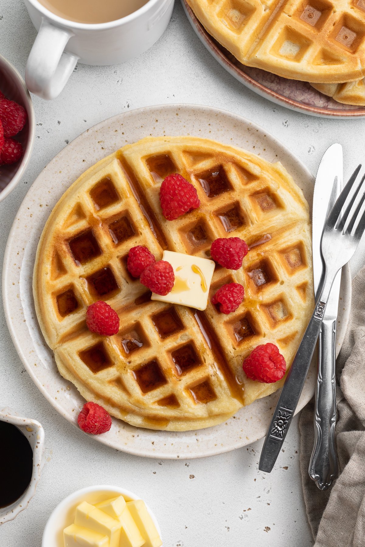 Overhead view of a plate of round, fluffy, Belgian-style gluten-free waffles topped with butter, syrup, and fresh red raspberries with silverware.