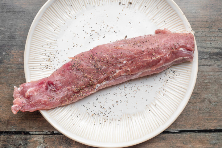 Overhead view of uncooked pork tenderloin, seasoned with salt and black pepper, resting on a white plate on a wooden table.