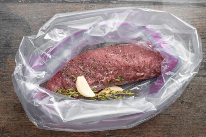 Overhead view of seasoned, uncooked pork tenderloin in a gallon ziploc bag with fresh herbs and smashed garlic.