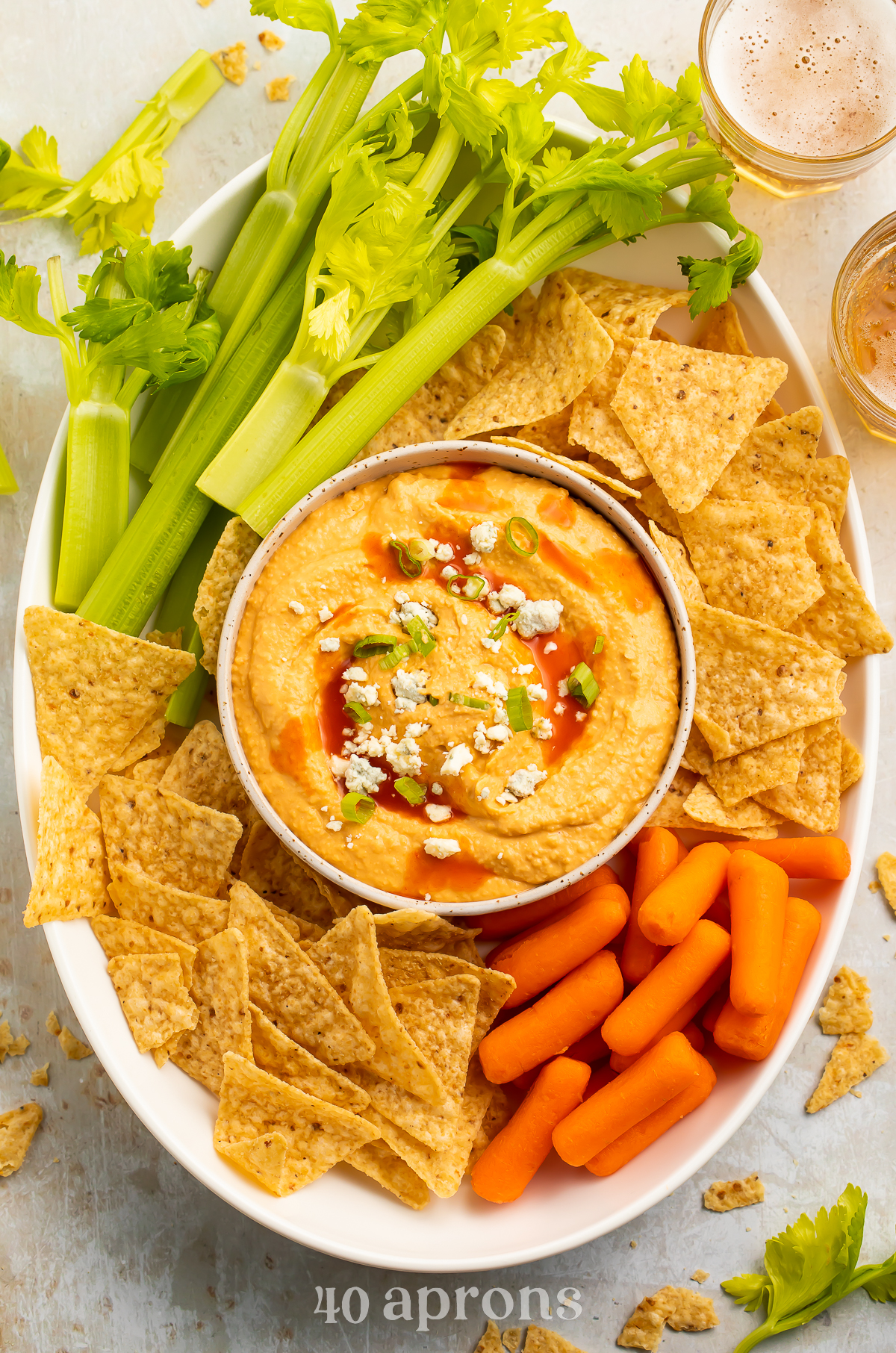 Overhead view of a bowl of buffalo hummus topped with blue cheese crumbles in the center of a platter surrounded by celery, carrots, and tortilla chips.