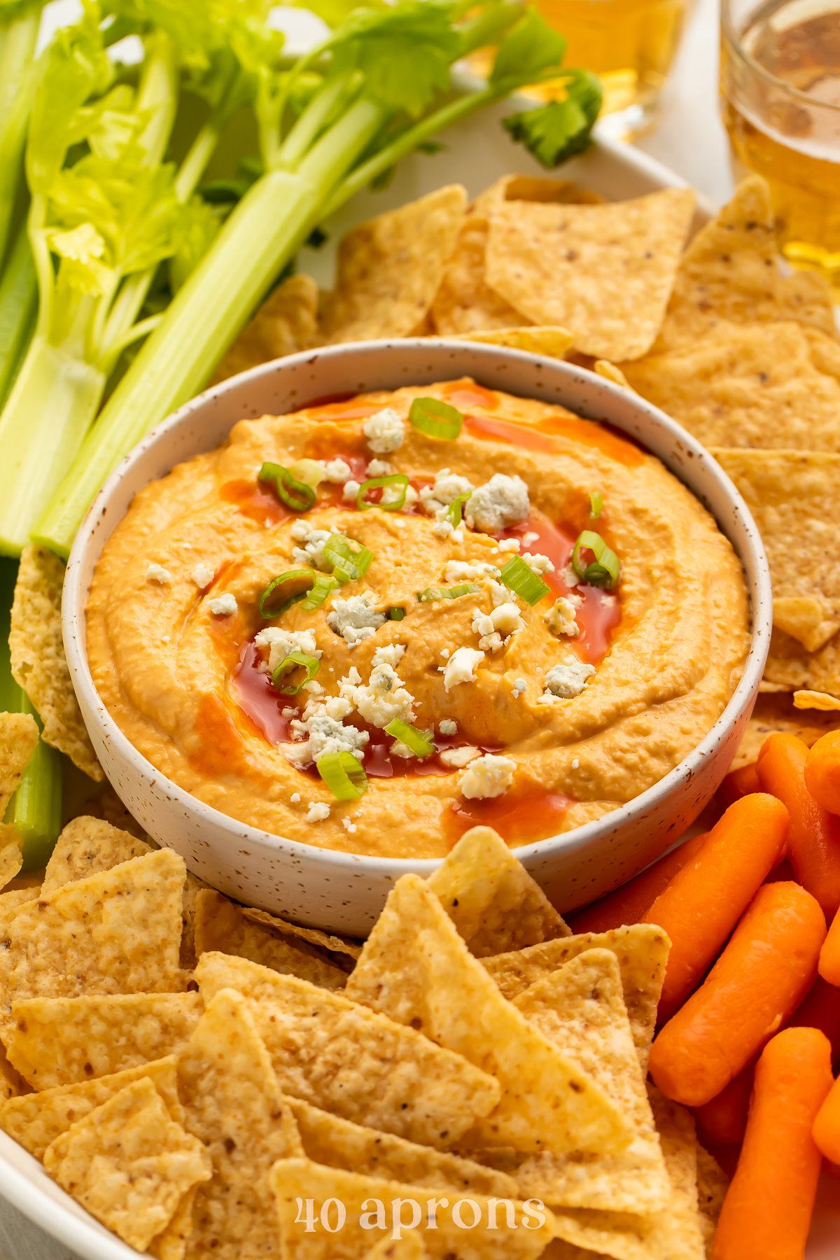 Angled, overhead view of buffalo hummus in a bowl, surrounded by carrot sticks, celery, and tortilla chips.