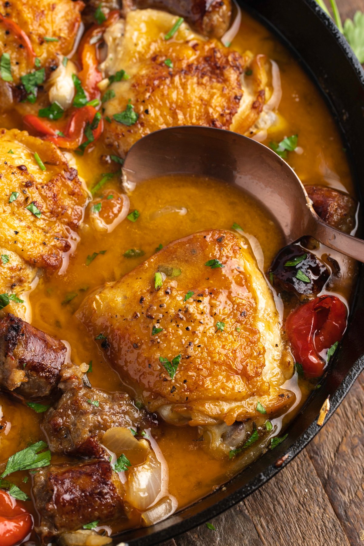 Overhead view of chicken scarpariello in a large cast iron skillet with a spoon on a wooden background.