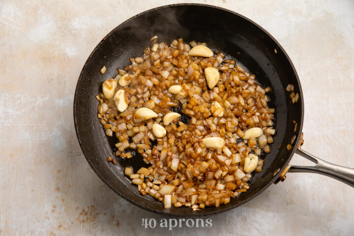 Overhead view of a cast iron skillet containing translucent sautéed onions, whole garlic cloves, and smashed garlic cloves.