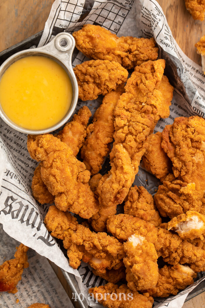 Close up overhead view of a pan lined with newspaper filled with chicken tenders and a small bowl of yellow honey mustard dipping sauce.