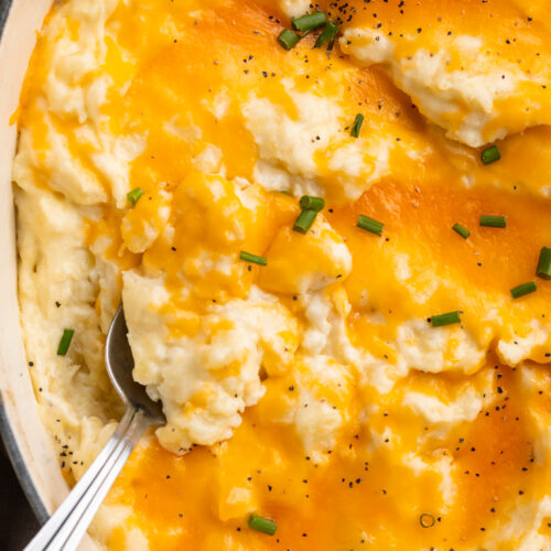 Overhead view of a large dish of cheesy mashed potatoes. A large silver spoon rests in the potatoes.