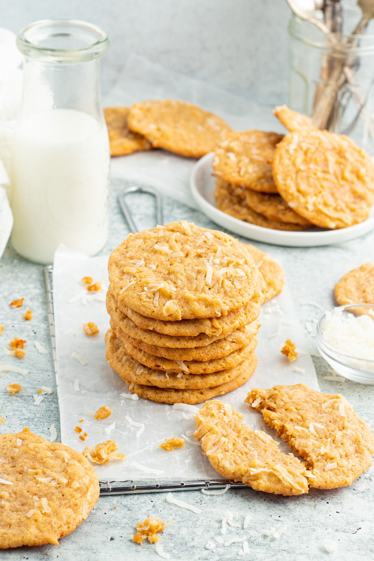 Angled, zoomed out view of a stack of coconut cookies next to a broken cookie and a glass of milk.