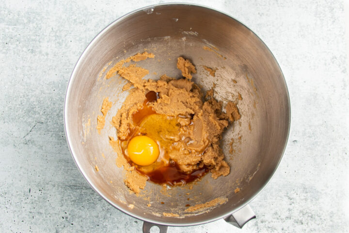 Overhead view of creamed butter and sugars with egg and vanilla extract in a large silver mixing bowl.