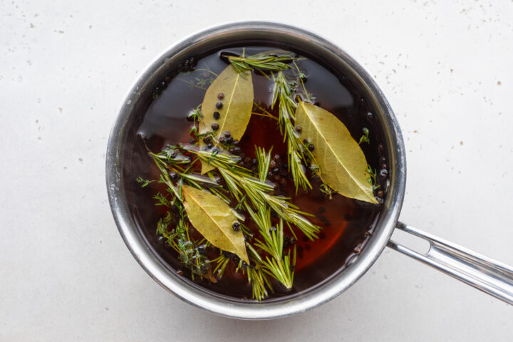 Overhead view of a quick turkey brine with brown sugar and sprigs of fresh herbs in a silver saucepan on a white tabletop.