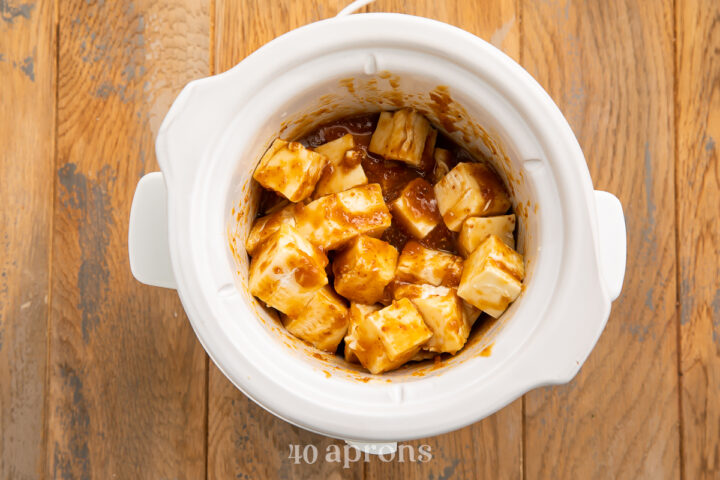 Overhead view of cubed brie cheese in a small white Crockpot with apricot preserves and apple juice.