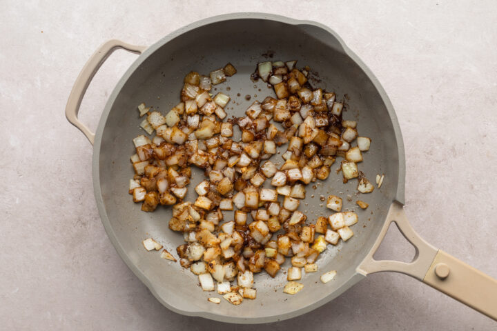 Overhead view of chopped onion cooking in rendered beef fat in a large skillet.