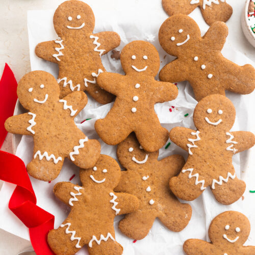 Overhead photo of gluten-free gingerbread men decorated with white cookie icing on a white table with red ribbon and holiday sprinkles.