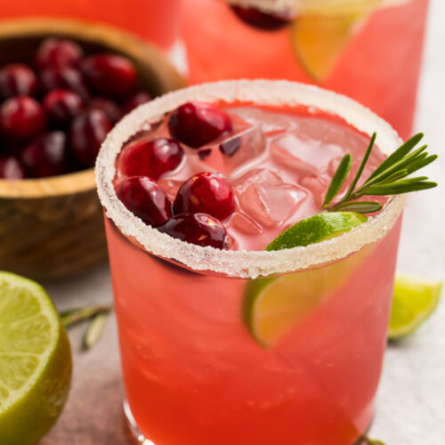 Bright red cranberry margaritas shown in stemless glasses rimmed with salt. Drinks are garnished with cranberries, rosemary, and lime wedges.