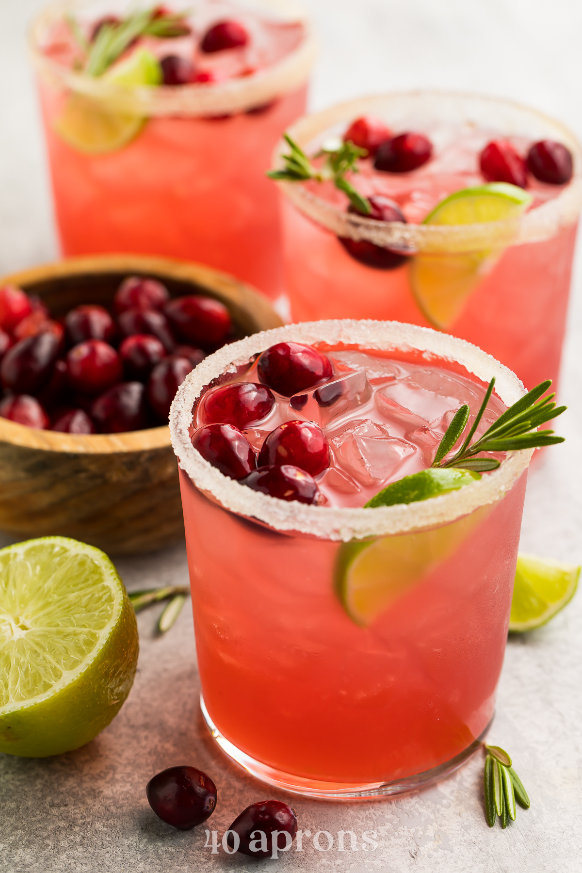 Bright red cranberry margaritas shown in stemless glasses rimmed with salt. Drinks are garnished with cranberries, rosemary, and lime wedges.