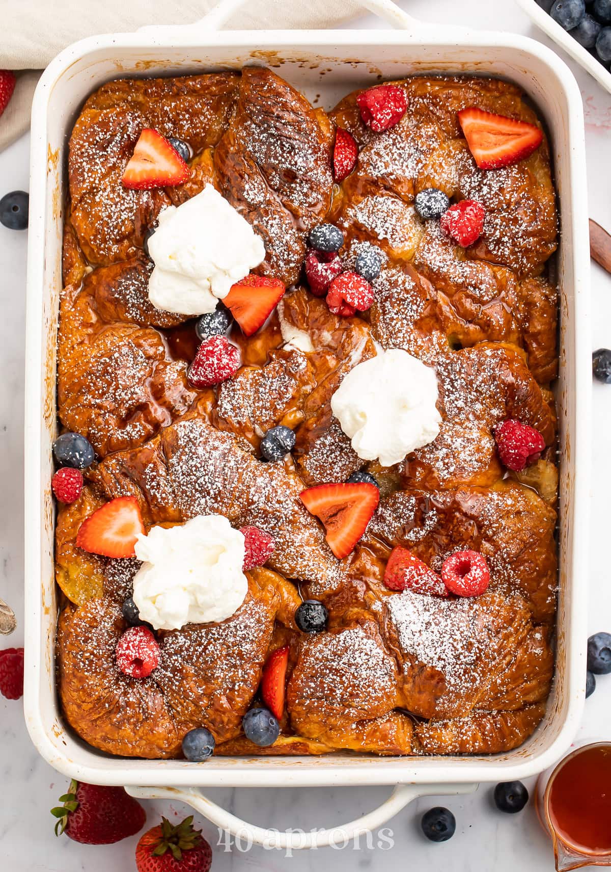 A large casserole dish holding a gorgeous French toast croissant casserole topped with powdered sugar, fresh fruit, and whipped cream.