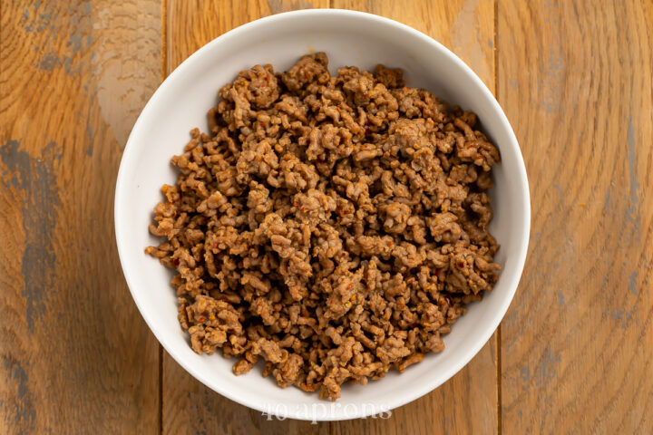 Overhead view of cooked and crumbled Italian sausage in a medium white bowl on a wooden countertop.