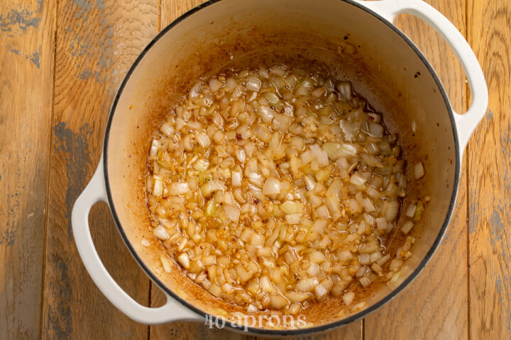 Overhead view of sautéed, translucent onions and minced garlic in rendered bacon fat in a large dutch oven.