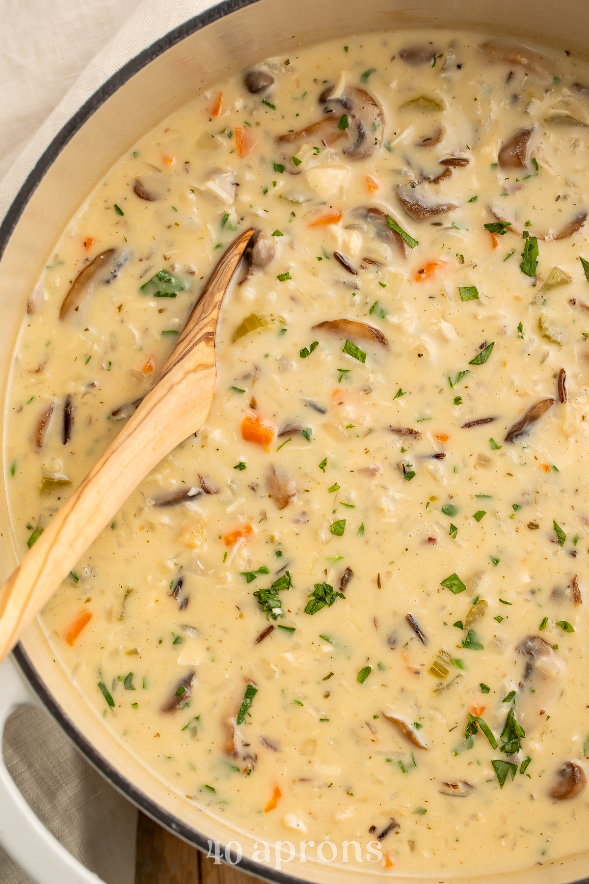 Overhead view of a large heavy pot containing creamy chicken and wild rice soup with mushrooms and carrots. A wooden spoon rests in the soup.