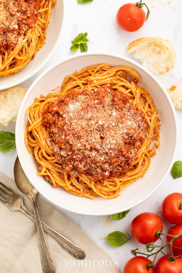 Overhead view of spaghetti noodles coated in rich, red Instant Pot spaghetti sauce in a large white pasta bowl on a table with tomatoes and silverware.