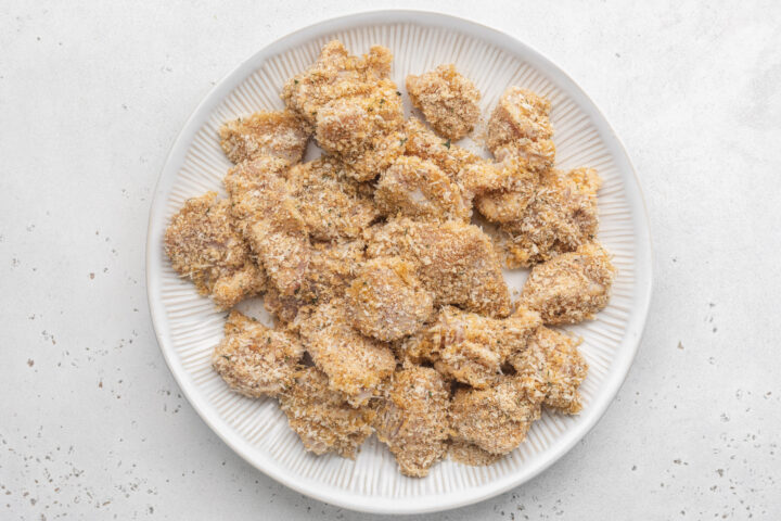 Breaded boneless, skinless chicken breast chunks on a large white plate ahead of being skewered.