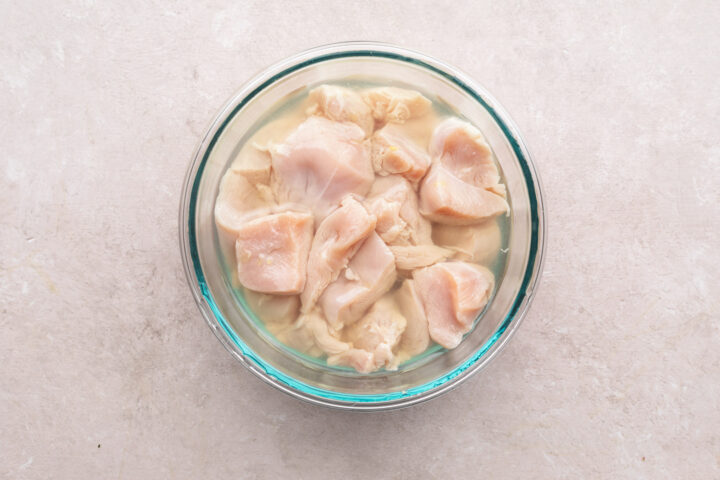 Bite-sized pieces of chicken breasts in a glass mixing bowl filled with pickle juice.