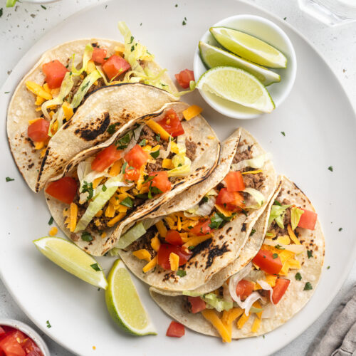 4 keto tacos in homemade keto tortillas lined up on a round white plate with lime wedges and toppings around them.