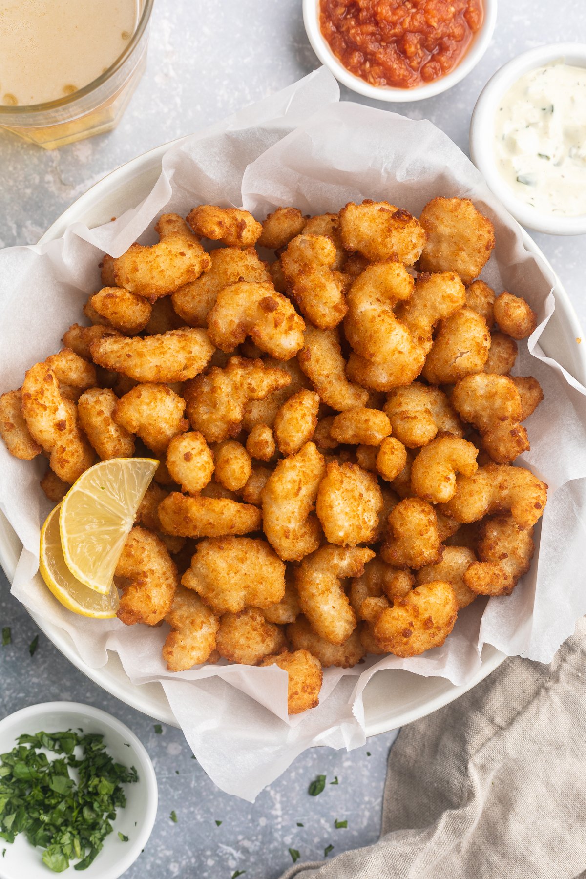 A large white bowl filled to the brim with golden, crispy popcorn shrimp cooked in the air fryer, with a lemon wedge to garnish and ramekins of dipping sauce surrounding.