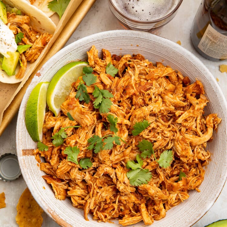 Instant Pot salsa chicken, shredded and transferred to a white serving bowl, topped with limes next to a pan of tacos.