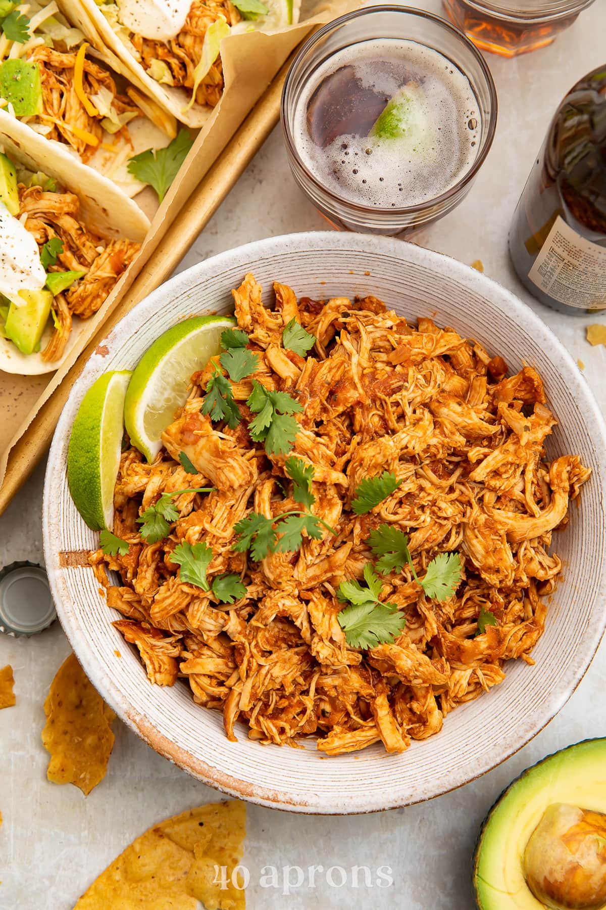 Instant Pot salsa chicken, shredded and transferred to a white serving bowl, topped with limes next to a pan of tacos.