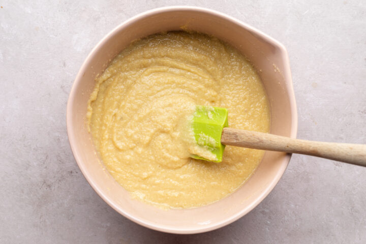 Keto donut batter in a large pink mixing bowl with a pour spout. A lime green silicone spatula with a wooden handle rests in the batter.