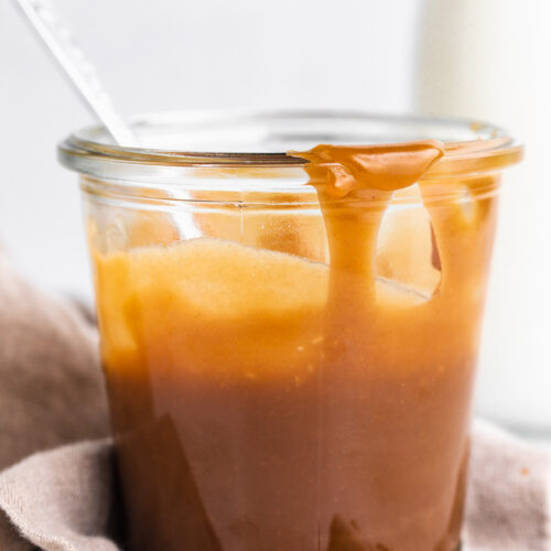 Side view of a glass jar of keto caramel sauce in front of a white background with a neutral napkin around the jar base.