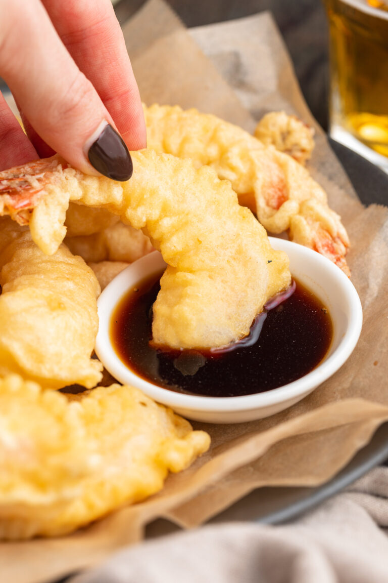 A large piece of shrimp tempura being dipped in a small bowl of soy sauce by a white woman's hand with black painted fingernails.