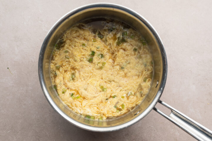 Scrambled eggs added to wonton egg drop soup broth in silver saucepan.