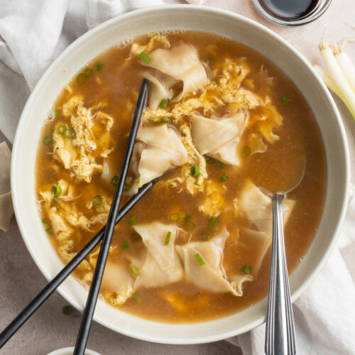 Top-down, overhead photo of a bowl of wonton egg drop soup on a neutral dining table surrounded by garnishes.