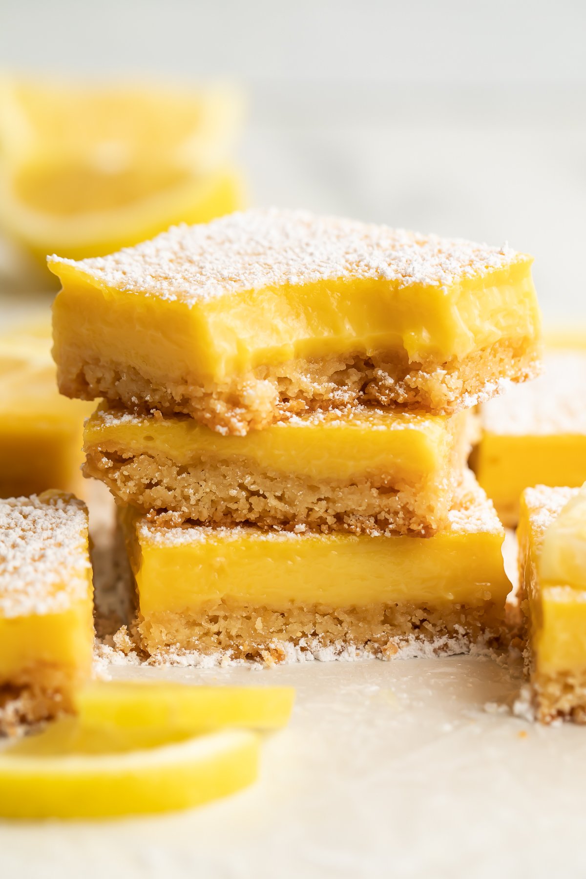 Three paleo lemon bars stacked on top of each other. A bite is missing from the corner of the lemon bar on top. Other lemon bars surround the stack, just out of focus.