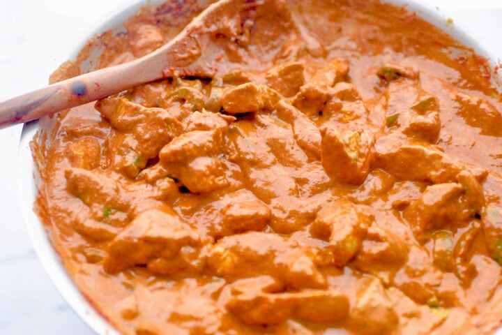 Rich, dark orange chicken tikka masala in a silver skillet with a wooden spoon.