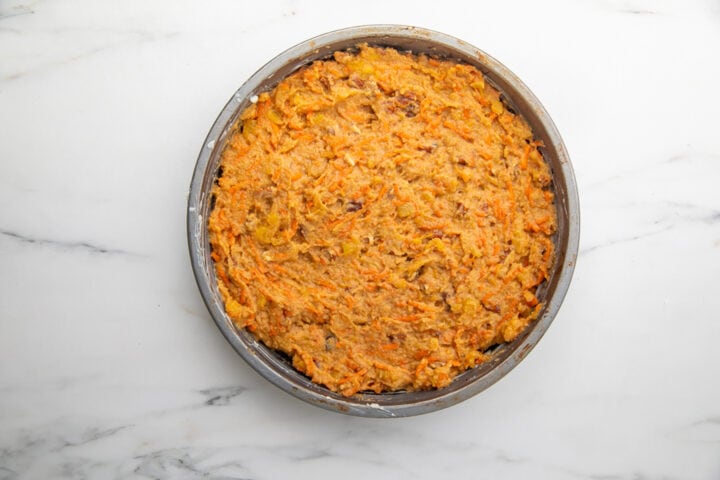 Prepared carrot cake batter transferred to a greased round cake pan on a marble countertop.