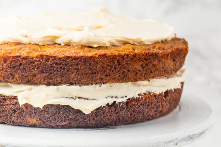 2 layers of carrot cake with cream cheese frosting on a white cake stand.