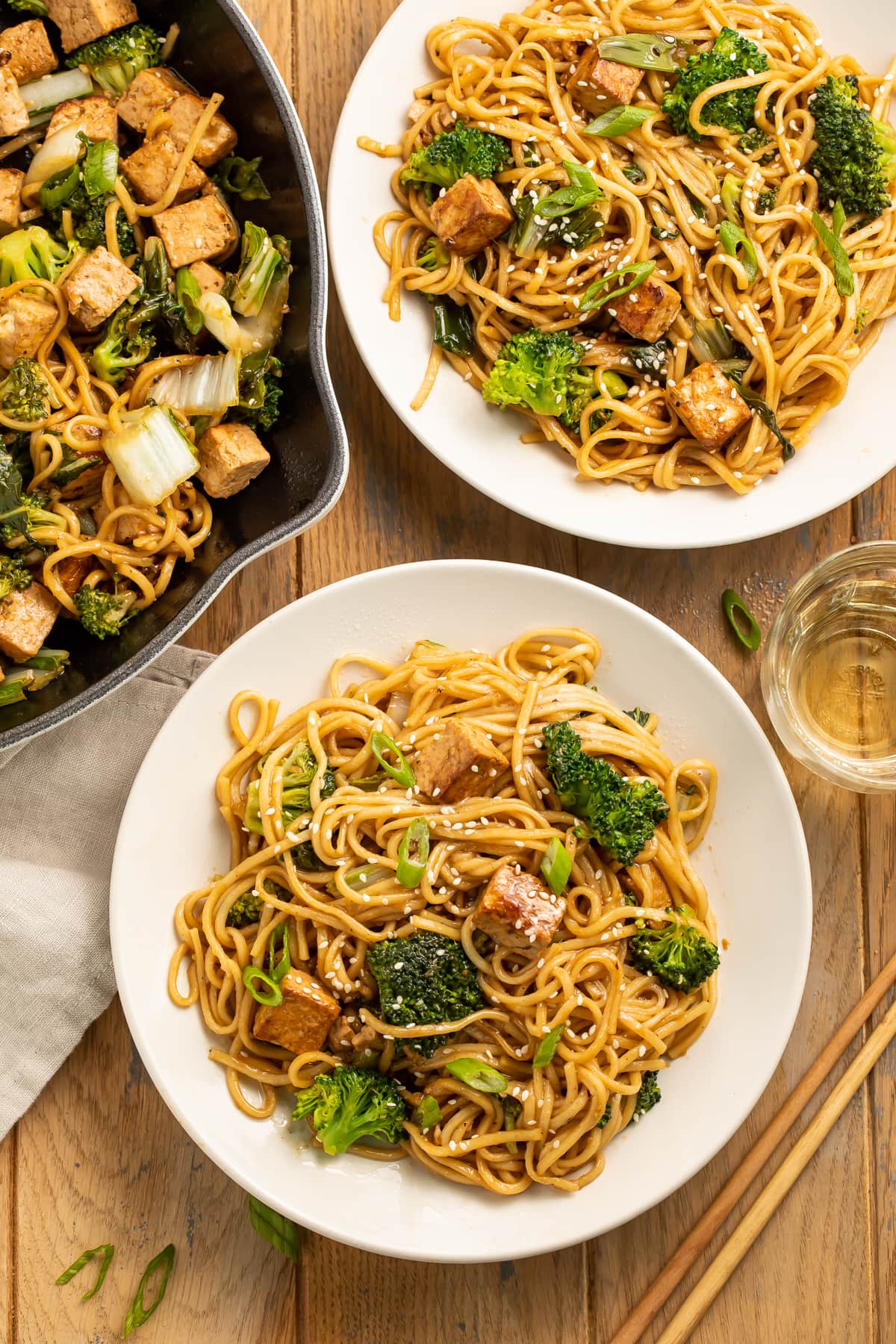 Overhead view of two large white plates piled with stir fry noodles and tofu next to a skillet of noodles.